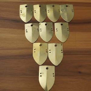 May include: A collection of ten brass-colored, shield-shaped metal pieces. Each piece has a rounded bottom, a pointed top, and a small hole near the top. Two or three small holes are near the bottom. The pieces are arranged on a wooden surface.