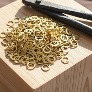 May include: A wooden block with a pile of small, gold-colored metal rings and a pair of black metal pliers. The rings are scattered across the top of the block, with some rings scattered around the edges.