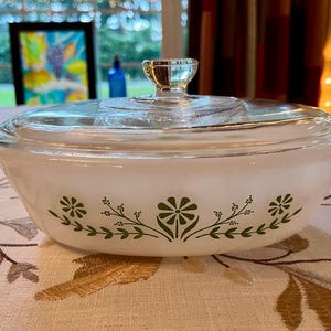 May include: A vintage white casserole dish with a clear glass lid. The dish features a green floral design around the rim. The lid has a silver-colored handle. This dish is perfect for serving and baking.