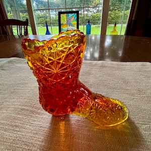 May include: An amber-colored glass boot with a detailed, cut-glass pattern. The boot transitions from a vibrant orange to a golden yellow. The decorative piece is likely vintage and sits on a neutral-colored fabric.