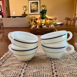 May include: Four white teacups with a teal blue stripe design. Each cup has a rounded shape with a handle. The cups are stacked in pairs on a brown woven placemat.