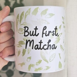 Peut inclure: Mug en c&eacute;ramique blanche avec l'inscription "But first, Matcha" en &eacute;criture noire. La tasse est d&eacute;cor&eacute;e de motifs de feuilles vertes. La tasse a une anse blanche.