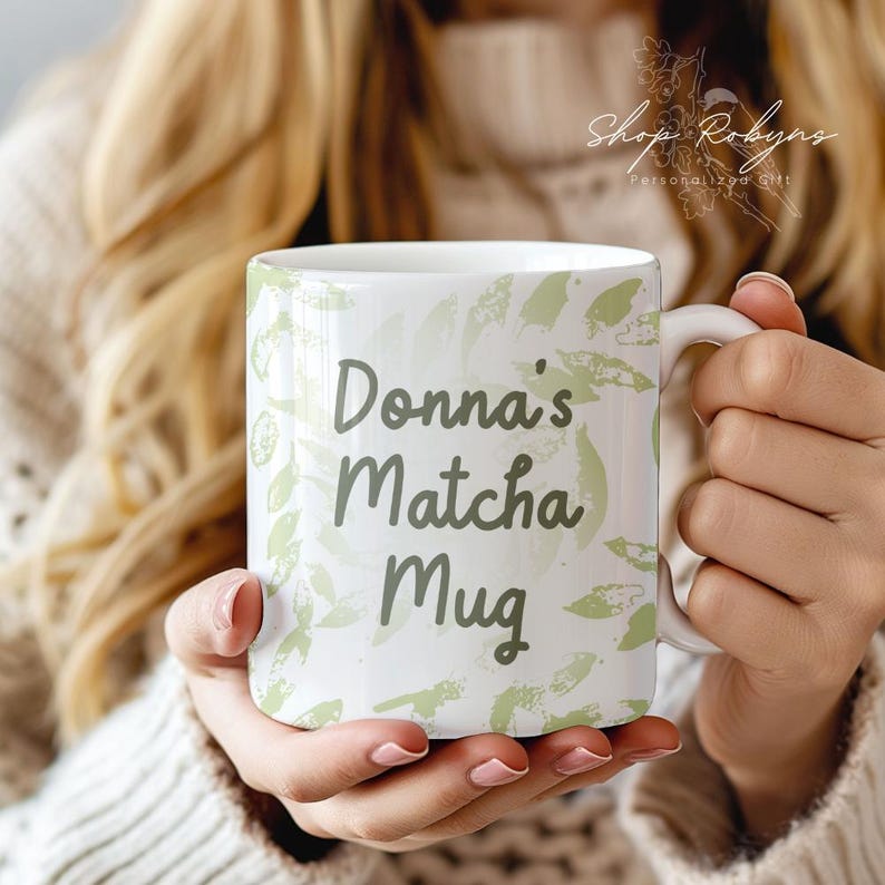 Peut inclure: Mug en c&eacute;ramique blanche avec l'inscription "Donna's Matcha Mug" dans un style manuscrit. La tasse est d&eacute;cor&eacute;e de motifs en forme de feuilles vert clair. La tasse a une anse blanche.