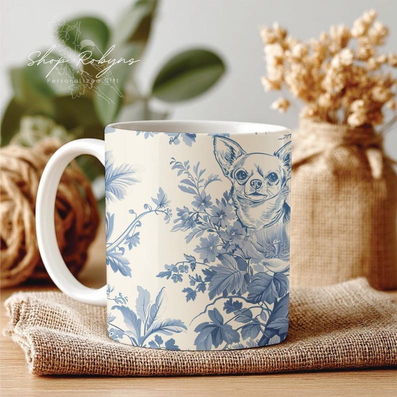 Pu&ograve; includere: Tazza in ceramica bianca con un motivo floreale blu e un'illustrazione di Chihuahua. La tazza ha un manico bianco e il testo "Shop Robyn's Personalized Gift". La tazza &egrave; su un panno di juta.