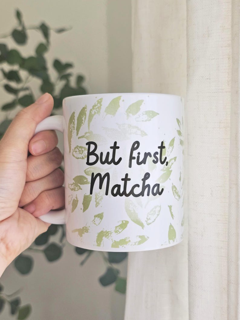 Peut inclure: Mug en c&eacute;ramique blanche avec l'inscription "But first, Matcha" en &eacute;criture noire. La tasse est d&eacute;cor&eacute;e de motifs de feuilles vertes. La tasse a une anse blanche.