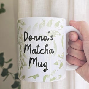Peut inclure: Mug en c&eacute;ramique blanche avec l'inscription "Donna's Matcha Mug" en &eacute;criture noire. La tasse pr&eacute;sente un motif de feuilles vertes et une anse blanche. La tasse est tenue dans une main.