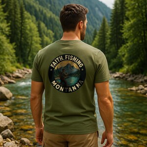 Puede incluir: Camiseta verde oliva con un gráfico circular en la espalda que muestra a un pescador, montañas y las palabras "Faith, Fishing, Montana". La camiseta de manga corta la lleva una persona de pie cerca de un río.