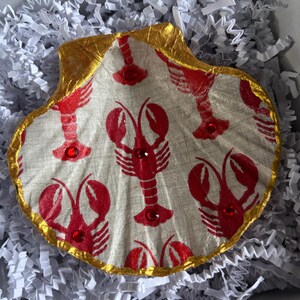 May include: A decorative scallop shell with a gold-painted rim and a fabric interior featuring a red lobster pattern. Red gemstones are placed on the lobsters. The shell is presented on white paper filler.