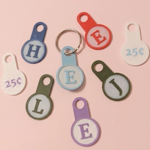 Könnte beinhalten: Eine Sammlung runder Kunststoffanhänger mit Löchern für Schlüsselringe. Die Anhänger zeigen die Buchstaben H, E, L, J und E in verschiedenen Farben wie Blau, Rot, Olivgrün und Lila. Zwei weiße Anhänger sind mit dem Text 25¢ bedruckt.