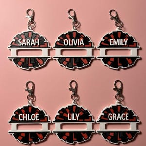 May include: Six circular keychains featuring a black and red pom-pom design with a white horizontal bar. Each keychain includes a silver metal lobster clasp and displays a unique name in white capital letters: Sarah, Olivia, Emily, Chloe, Lily, and Grace.