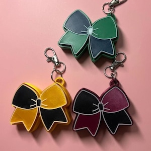 May include: Three bow-shaped keychains with silver-toned metal lobster clasps. One keychain features dark blue and light green, another is yellow and black, and the third is maroon and black. Each bow has a central knot and textured surfaces.