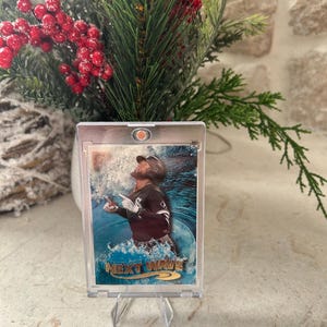 May include: A baseball card in a clear protective case, featuring a player in a white and black uniform amidst a blue water background. The card reads "NEXT WAVE" in a stylized font. The card is displayed on a clear stand.