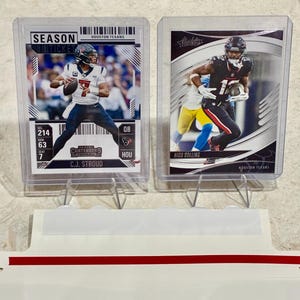 May include: Two football trading cards in protective sleeves. One card features C.J. Stroud in a white and blue uniform, with the text "SEASON TICKET" and "HOUSTON TEXANS." The other card shows Nico Collins in a black and red uniform. Both cards are displayed on a stand.