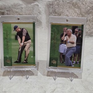 May include: Two golf trading cards in protective cases. One card shows a golfer in a black vest and cap swinging a club. The other card depicts a golfer in a light yellow shirt mid-swing, with spectators in the background. Both cards have a green and gold design.
