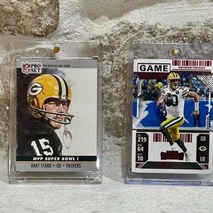 May include: Two NFL football trading cards in protective cases. One card features Bart Starr, MVP Super Bowl I, with the Packers logo. The other card shows a Green Bay Packers player in action, with game stats.
