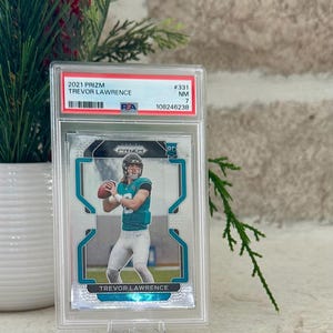 May include: A 2021 Prizm Trevor Lawrence football trading card, graded and encased in a protective holder. The card features a player in a teal and black uniform. The holder is displayed on a clear plastic stand, with text that reads "2021 Prizm Trevor Lawrence".