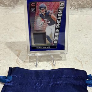 May include: A football trading card of Rome Odunze in a clear protective case. The card features a player in a Chicago Bears uniform with the text "ROOKIE PHENOM". A blue satin drawstring bag is in the foreground.