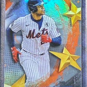 May include: A baseball trading card featuring Juan Soto in a New York Mets uniform. The card has a holographic background with gold star accents. The player is wearing a blue helmet and red gloves. The card is encased in a protective sleeve.