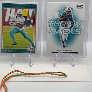 May include: Two Score trading cards featuring De'Von Achane of the Miami Dolphins in protective plastic cases, displayed on clear stands, accompanied by a thin, braided orange and green cord resting on the surface in front of the cards.