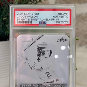 May include: A baseball card encased in a clear protective holder. The card features a black and white illustration of a baseball player and the text "BRIGHT & SHINY". The card is labeled "2024 LEAF VIVID JACOB WILSON".