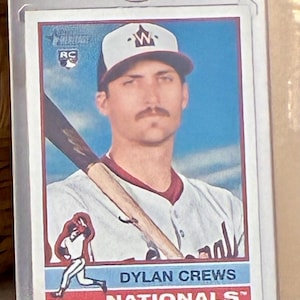 May include: A baseball card featuring Dylan Crews of the Nationals. The card shows Crews in a white and burgundy baseball uniform, holding a bat. The card is in a protective case. The text "DYLAN CREWS" and "NATIONALS" are visible.