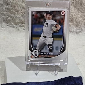 May include: A baseball card of Jackson Jobe of the Detroit Tigers in a protective case. The card features a photo of Jobe pitching in his team uniform. The card is displayed on a clear plastic stand.