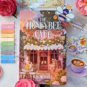 May include: A book cover titled "The Honeybee Cafe" with a cafe illustration, surrounded by stickers, sticky notes, and pink flowers. A bee-shaped sticker is on the book. The book club card reads "The Honeybee Cafe Book Club".