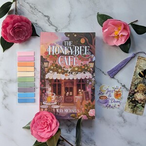 May include: A book titled "The Honeybee Cafe" with a colorful illustration of a cafe, surrounded by pink flowers, a bookmark with a bee and flowers, sticky notes, and a sticker with a cup of coffee.
