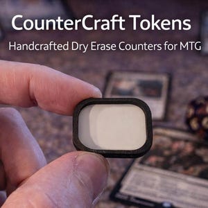 Peut inclure: Un jeton de comptoir effaçable à sec blanc avec un cadre noir est tenu entre les doigts. Le texte "CounterCraft Tokens" et "Handcrafted Dry Erase Counters for MTG" sont visibles. D'autres cartes de jeu et un dé sont en arrière-plan.