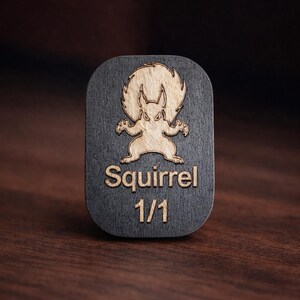 May include: A wooden plaque with a dark gray background and a light brown squirrel design. The word "Squirrel" and the number "1/1" are engraved in a light brown color. The plaque is rectangular with rounded corners.