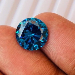 May include: A round, faceted gemstone with a brilliant blue hue. The gem is held between fingers, showcasing its clarity and the play of light across its many facets. The gemstone's color ranges from deep teal to a lighter, vibrant blue.
