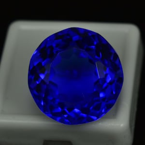 May include: A faceted, round, deep blue gemstone. The gem is set against a white background, with the facets reflecting light to create a brilliant display of color and sparkle. The gemstone's rich blue hue is the focal point.