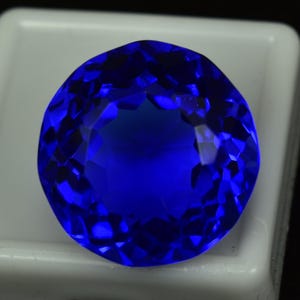 May include: A round, faceted, deep blue gemstone. The gem has a brilliant cut with many facets that reflect light, creating a sparkling effect. The stone is set against a white background.