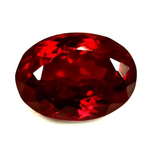 May include: A faceted, oval gemstone in a deep red colour. The surface of the gem reflects light, creating bright highlights. The stone is set against a white background, emphasising its colour and clarity. Suitable for jewellery.