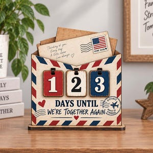 Puede incluir: Un calendario de cuenta regresiva de madera con forma de sobre y bordes a rayas rojas y azules. Incluye tres placas rectangulares colgantes con los números 1, 2 y 3. El texto dice: Days Until We're Together Again, con un sello postal.