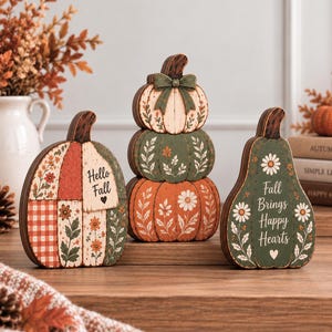 May include: Three decorative wooden pumpkin figures featuring painted floral patterns, plaid details, and text. One pumpkin reads Hello Fall, while another reads Fall Brings Happy Hearts. The set includes a stacked pumpkin design with a green bow.