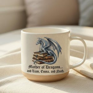 Op de afbeelding: Witte keramische mok met een illustratie van een zilveren en blauwe draak en de tekst "Mother of Dragons... and Liam, Emma, and Noah." De draak zit op boeken. De mok heeft een bruine basis.