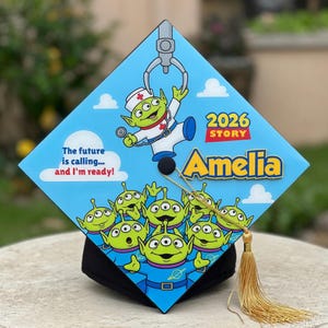 Op de afbeelding: Een blauwe afstudeerhoed met een cartoon alien ontwerp. De hoed heeft de tekst "2026 Story Amelia" en de zin "The future is calling... and I'm ready!" Een gouden kwast hangt aan de hoed.