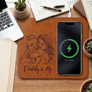 May include: A brown wireless charging pad with a laser-etched image of a father and child embracing, with the text "Daddy & Me." A smartphone charging wirelessly displays a green charging symbol and 67% charged.