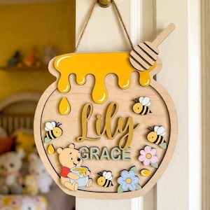 May include: A round wooden wall sign shaped like a honey pot with a honey dipper at the top. The sign features the name Lily Grace in script and block lettering, a cartoon bear, several small bees, flowers, and yellow honey drips.
