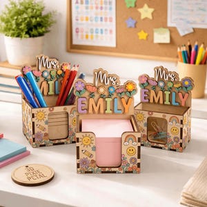 Può includere: Tre organizer da scrivania in legno con disegni floreali e arcobaleno. Ognuno presenta un nome, "Mrs. Emily" o "Mrs. Mil", e contiene penne, matite e carta per appunti. Gli organizer hanno un'estetica retrò e colorata.