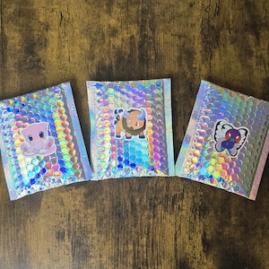 K&ouml;nnte beinhalten: Drei holografische Luftpolstertaschen mit Aufklebern von Zeichentrickfiguren. Die Umschl&auml;ge haben eine regenbogenfarbene, schillernde Oberfl&auml;che und sind versiegelt. Die Aufkleber zeigen ein rosa Wesen, einen braunen Bullen und einen blauen Schmetterling.