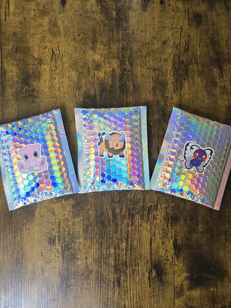 K&ouml;nnte beinhalten: Drei holografische Luftpolstertaschen mit Aufklebern von Zeichentrickfiguren. Die Umschl&auml;ge haben eine regenbogenfarbene, schillernde Oberfl&auml;che und sind versiegelt. Die Aufkleber zeigen ein rosa Wesen, einen braunen Bullen und einen blauen Schmetterling.