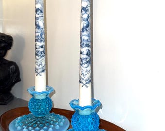 May include: Two white taper candles with blue and white illustrations sit in matching turquoise glass candle holders. The candle holders have a textured, hobnail design and sit on a wooden tray.