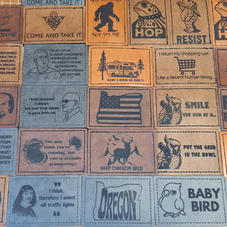 May include: A collection of rectangular leather patches in various colors, each with unique designs and text. The patches feature images of a bigfoot, a frog, an owl, and text like "COME AND TAKE IT" and "OREGON".