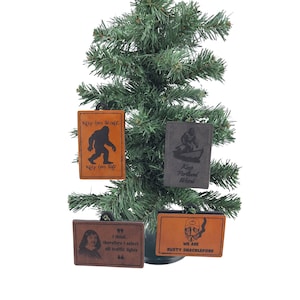 Op de afbeelding: Een kleine kunstkerstboom versierd met vier leren ornamenten. De ornamenten zijn rechthoekig met verschillende ontwerpen en tekst, waaronder een Bigfoot-silhouet en de zinnen "Keep him Safe" en "Keep Portland Weird."