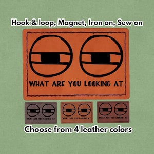 Può includere: Toppa in pelle arancione con due forme di occhi neri e la scritta "WHAT ARE YOU LOOKING AT". La toppa è disponibile in quattro colori di pelle e può essere applicata con velcro, magnete, termoadesiva o cucita.