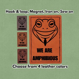 May include: An orange rectangular patch featuring a smiling frog face and the text "WE ARE AMPHIBIOUS" in black. Three smaller patches with the same frog design and text are to the left. The text "Hook & loop, Magnet, Iron on, Sew on" is at the top.