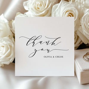 Pode incluir: Um cartão branco com as palavras "thank you" em elegante escrita preta, com "Olivia & Chloe" abaixo. O cartão é colocado sobre um fundo de rosas brancas e uma pequena caixa com anéis de ouro.