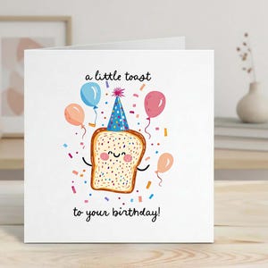 May include: A white birthday card with a cartoon illustration of a slice of toast wearing a party hat and surrounded by balloons and confetti. The text reads "a little toast to your birthday!"
